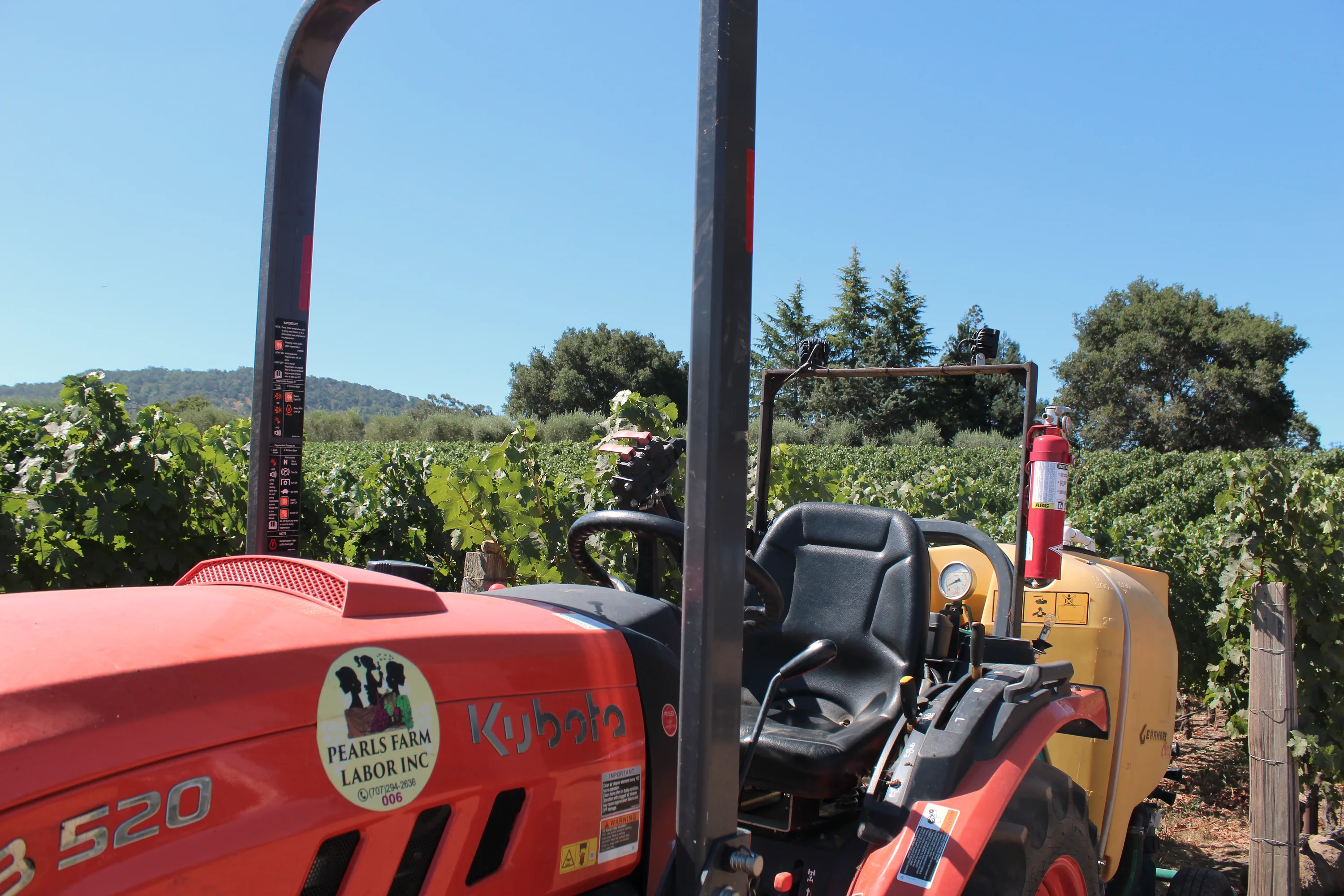 Red tractor with a yellow sprayer tank beside vineyard rows